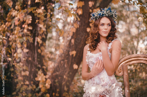 Beautiful brown-haired woman standing in a flower wreath on his head and a beautiful white dress standing in a forest leaning against a harp