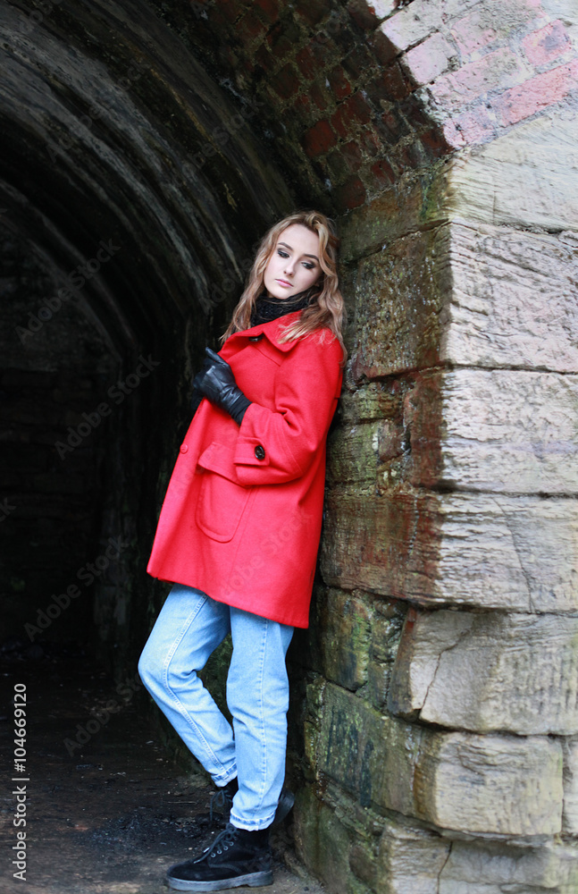 Woman wearing a red coat leaning against an old wall of a tunnel