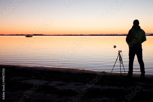 Silhouette of tall nature photographer at tripod taking picture on beach at sunset