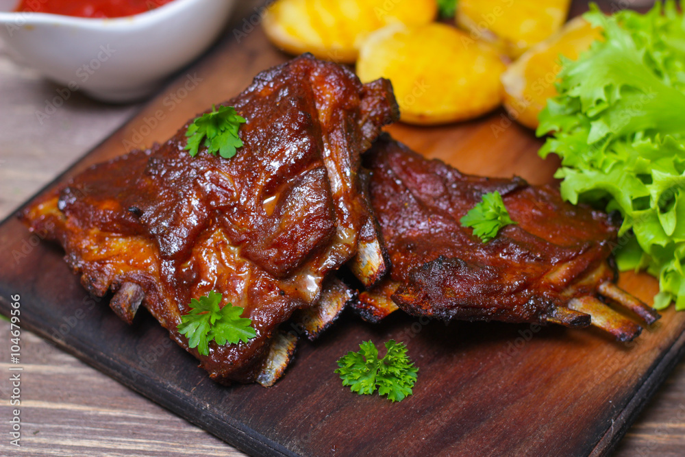 Appetizing rosy and barbecued lamb ribs seasoned with a barbecue sauce and served with fresh herbs and potatoes on an old rustic wooden chopping board Appetizing rosy and barbecued lamb ribs seasoned with a barbecue sauce and served with fresh herbs and potatoes on an old rustic wooden chopping board
