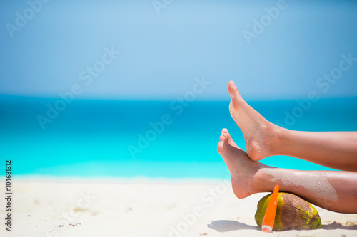 Female tanned smooth legs with suncream and coconut on white beach