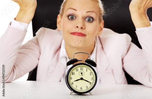 Surprised business woman lsitting behind the desk with alarm clock photo