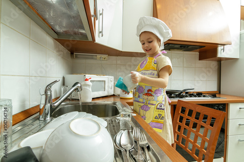 Obraz na plátně Little girl in white chef hat is washing dishes in the kitchen.