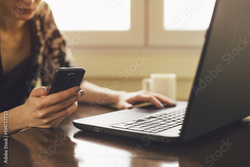 Unrecognizable young woman looking at smartphone while using laptop photo