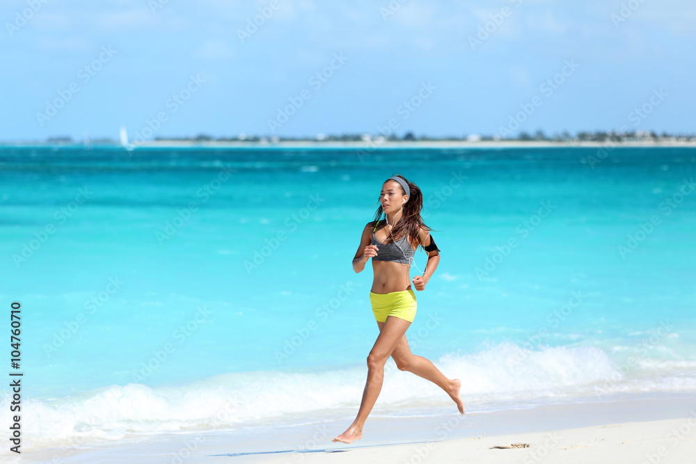 Female runner running on beach - summer vacations exercise. Asian female athlete strength training during holiday travel working out her cardio listening to music on her smartphone with sport armband.