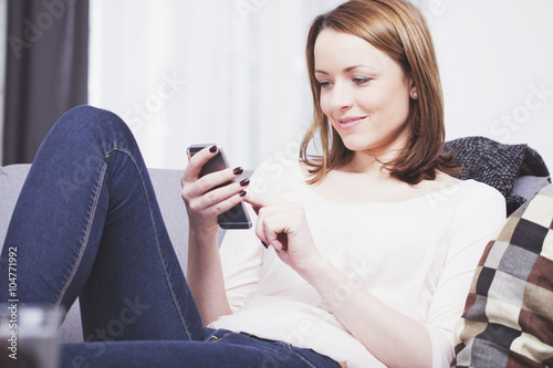 Pretty brown haired young woman dressed casual sitting on a sofa while playing with her smartphone