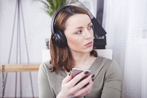 Serious looking brown haired girl with headphones holding smartphone looking pensively to the side
