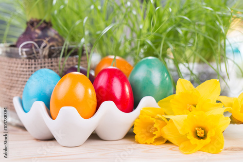 Wielkanocne dekoracje z kolorowymi jajami wielkanocnymi pisankami w ceramicznym stojaku, żonkilami i zielonymi roślinami w tle