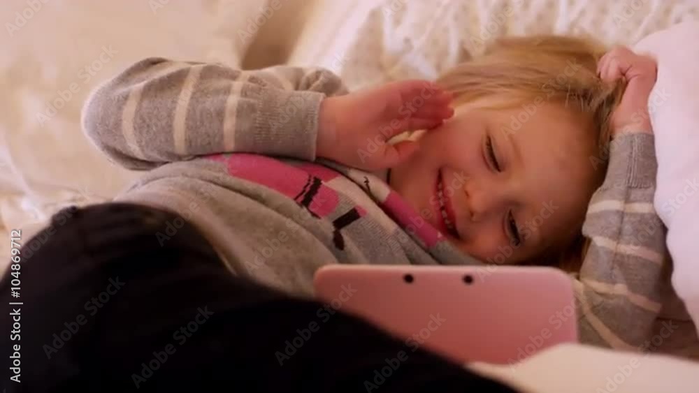 A little girl laying on her bed watching a show on her handheld gaming system, and she smiles and waves A little girl laying on her bed watching a show on her handheld gaming system, and she smiles and waves