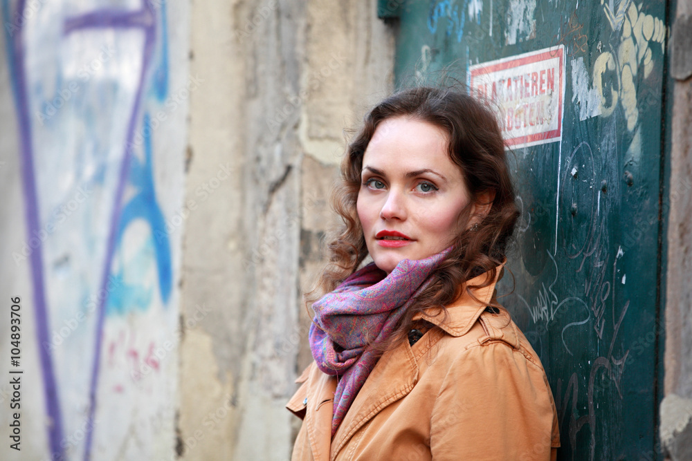 Beautiful brunette woman with beige coat standings at a house with graffiti