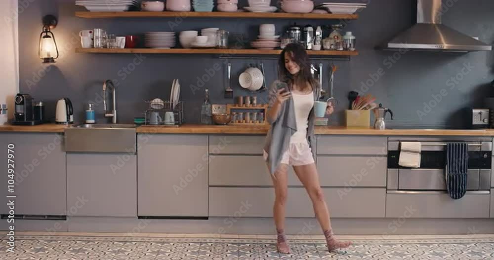 Happy young woman dancing in kitchen wearing pajamas smart phone coffee morning