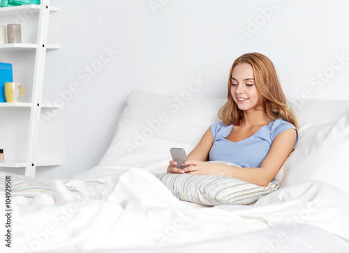 happy woman or girl with smartphone in bed at home