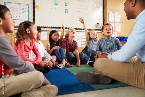 Elementary school kids and teacher sit cross legged on floor