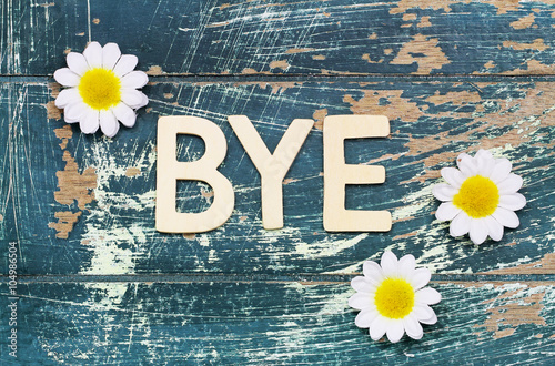 Word bye written with wooden letters on rustic surface and white daisies
