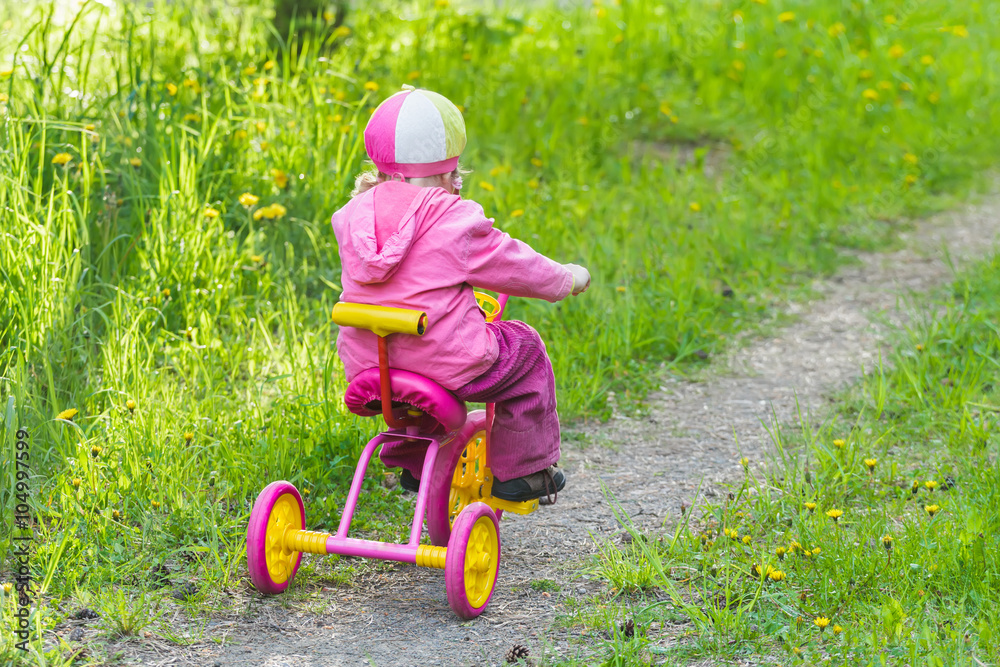 Back view full length portrait of little girl riding kids pink and yellow tricycle on park track Back view full length portrait of little girl riding kids pink and yellow tricycle on park track