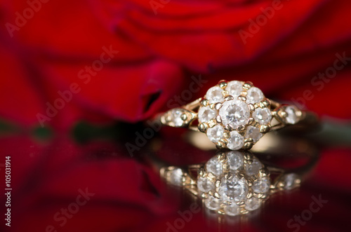 Gold ring with diamond on a red background. Ring is reflected on the mirror surface. In the background rose, which is out of focus and is reflected on the mirror surface.
