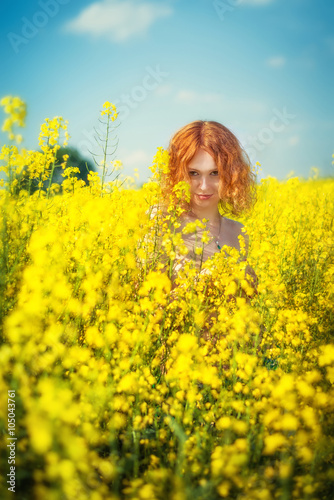 Smiling red-haired pretty girl looks out of the yellow flowers on the field 