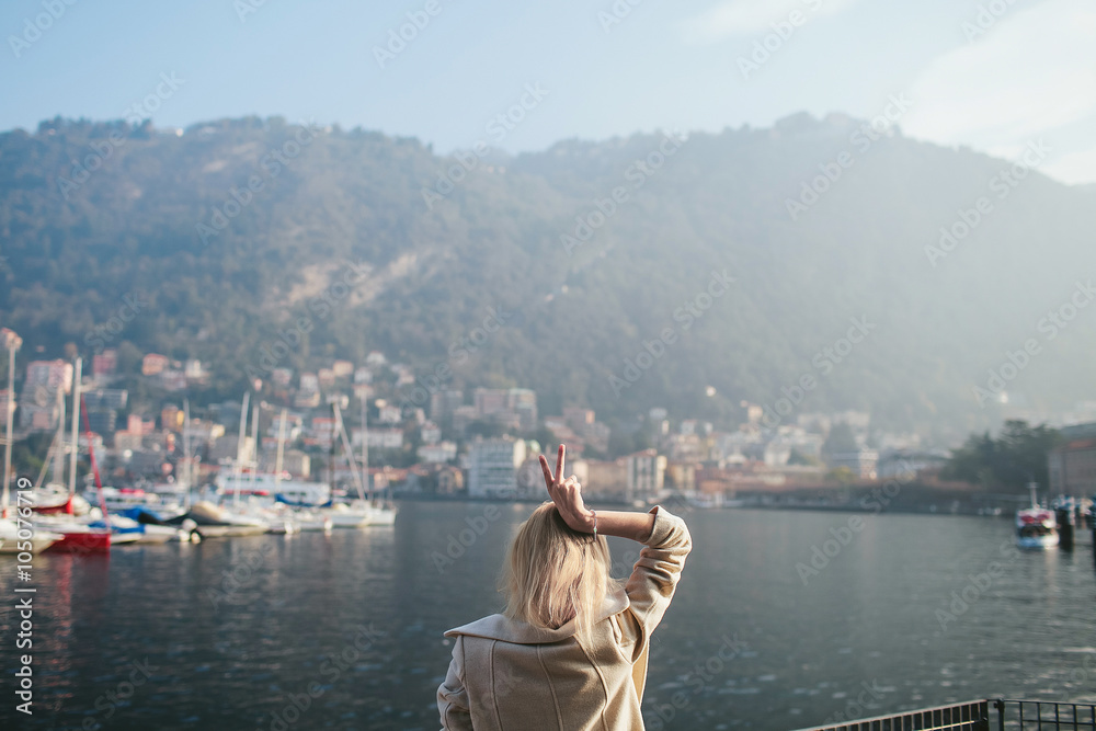 blonde girl stands with his back on the waterfront with views of the mountain lake of Como