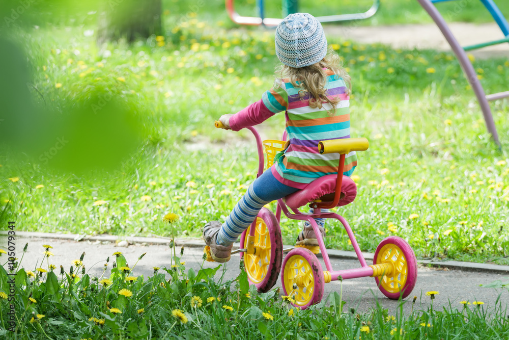 Back view full length portrait of preschooler girl riding kids pink and yellow tricycle on playground track Back view full length portrait of preschooler girl riding kids pink and yellow tricycle on playground track