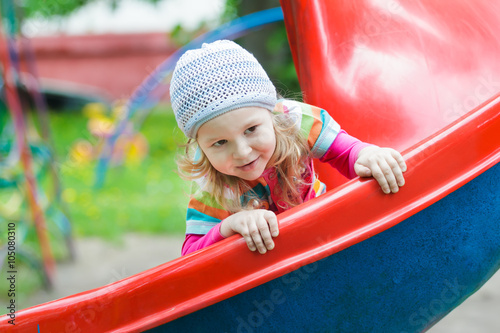 Smiling little blonde girl sliding down red plastic playground slide outdoors on spring