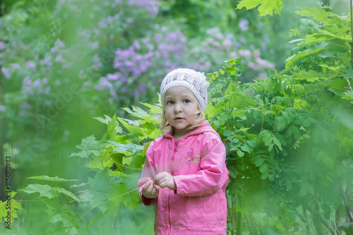 Two years old cute girl wearing white knitted hat at green spring garden lilac shrubbery background