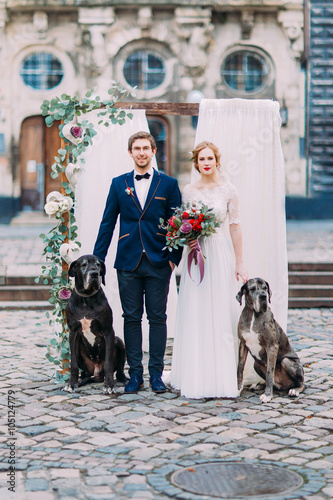 Stylish young wedding couple with two purebread dogs posing with wedding decorations on the baroque architecture on background photo