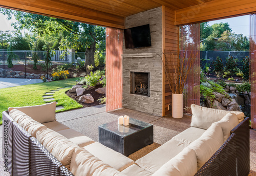 Beautiful covered patio outside new luxury home with television, fireplace, and lush green yard