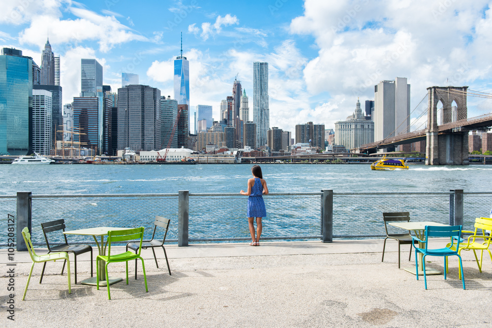 Naklejka premium New York city skyline waterfront lifestyle - American people walking enjoying view of Manhattan over the Hudson river from the Brooklyn side. NYC cityscape with a public boardwalk with tables.