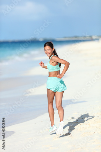 Happy healthy lifestyle woman runner jogging on white sand looking back smiling at camera. Asian fitness athlete young adult running on vacation beach showing off fit body and toned thighs and butt.