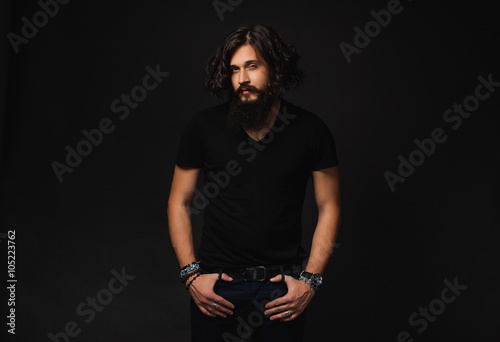 man in a black shirt and pants with a beard and mustache posing in the studio on a black background, the macho male muscular guy,handsome man portrait of a brutal male movie star fashionable clothes