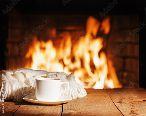 Fototapeta White cup of tea or coffee and woolen scarf near fireplace.
