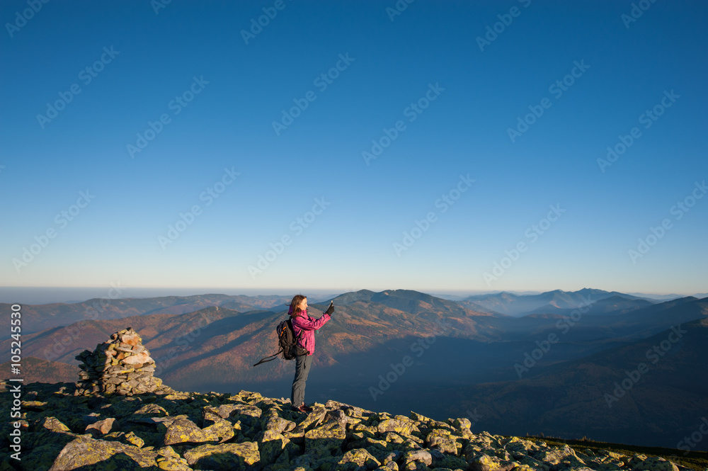 Young beautiful girl standing on rocky mountain top and taking picture with her smartphone. Wide angle view. Ecotourism and healthy lifestyle concept. Sunny fall day.