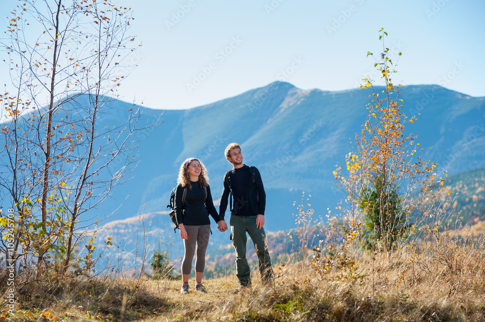 Young happy couple tourists holding hands smiling and looking into distance on a warm sunny autumn day in the mountains. Mountain on the background.