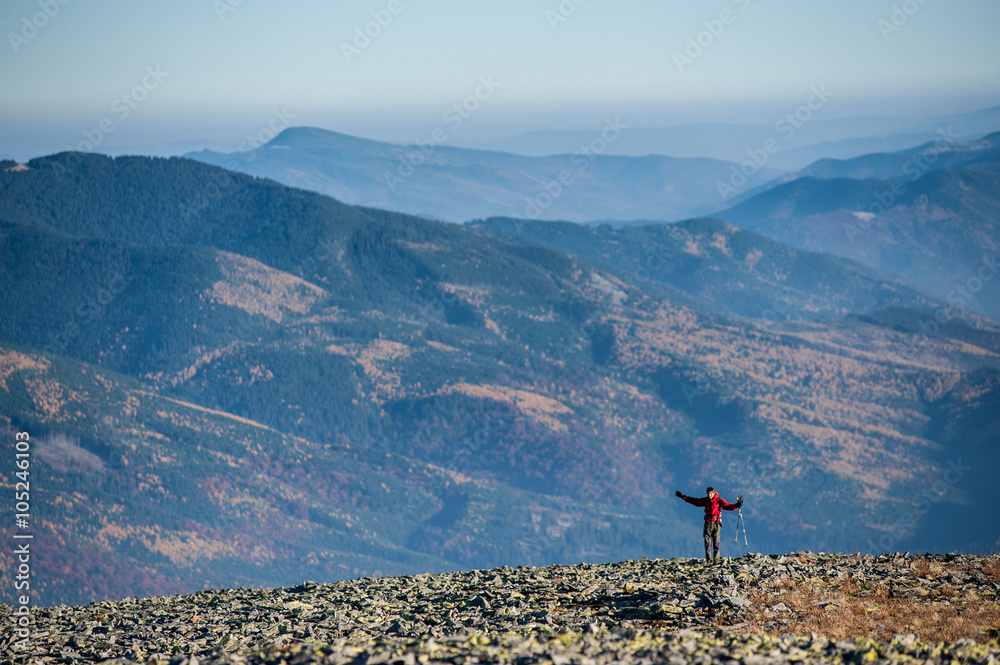 Fototapeta premium Happy hiker/backpacker standing on rocky mountain plato with amazing mountains on background. Man raised arms, success gesture, holding tripod. Ecotourism and healthy lifestyle concept. Copy space.