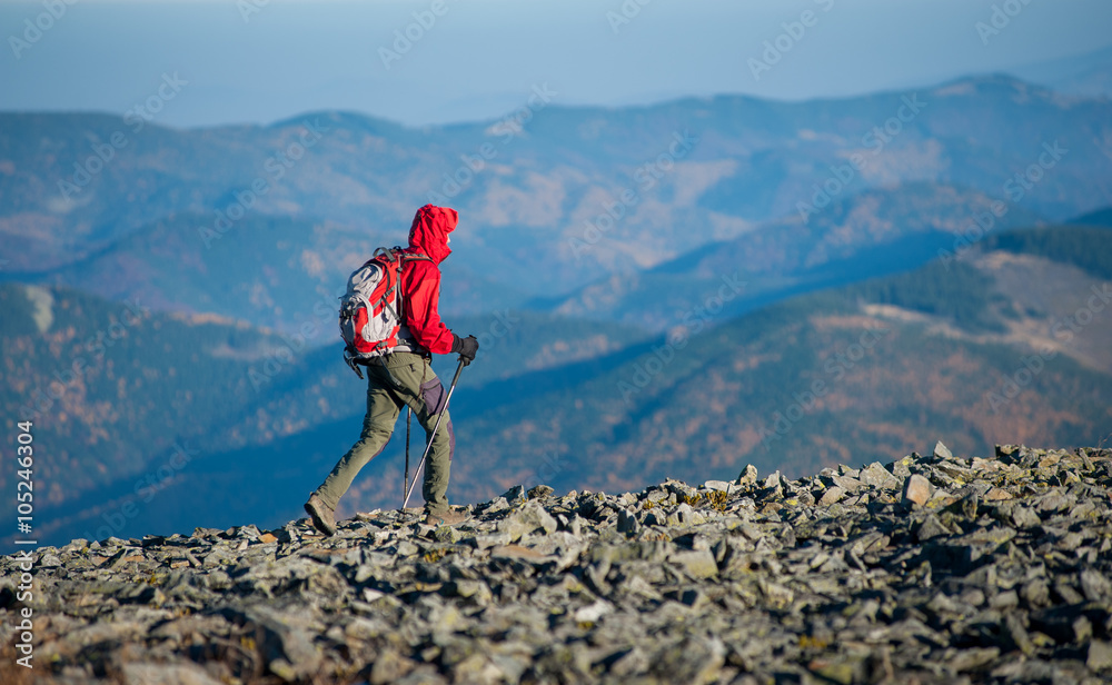 Male hiker backpaker walking on the rocky mountain ridge with beautiful mountains on background. Man is wearing red jacket and has trekking sticks and backpack on. Sunny fall day.
