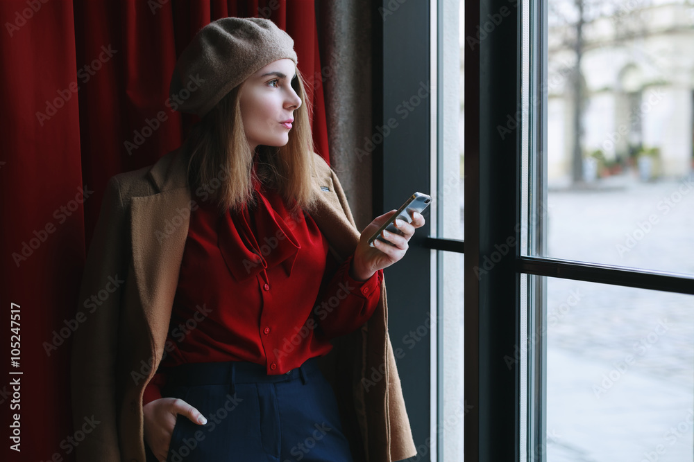 Portrait of  young beautiful woman wearing stylish classic clothes holding mobile phone and looking through the window. Close up. 
