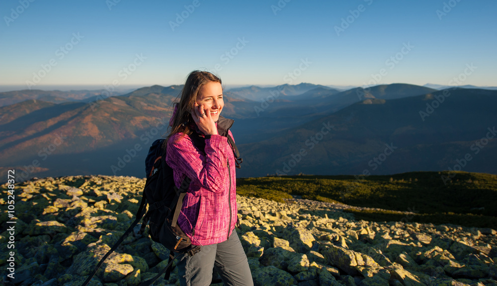 Naklejka premium Portrait of young happy woman backpacker standing on rocky top of the mountain, smiling, looking to the sun and talking cell phone. Side view. Healthy lifestyle concept. Sunny fall evening.