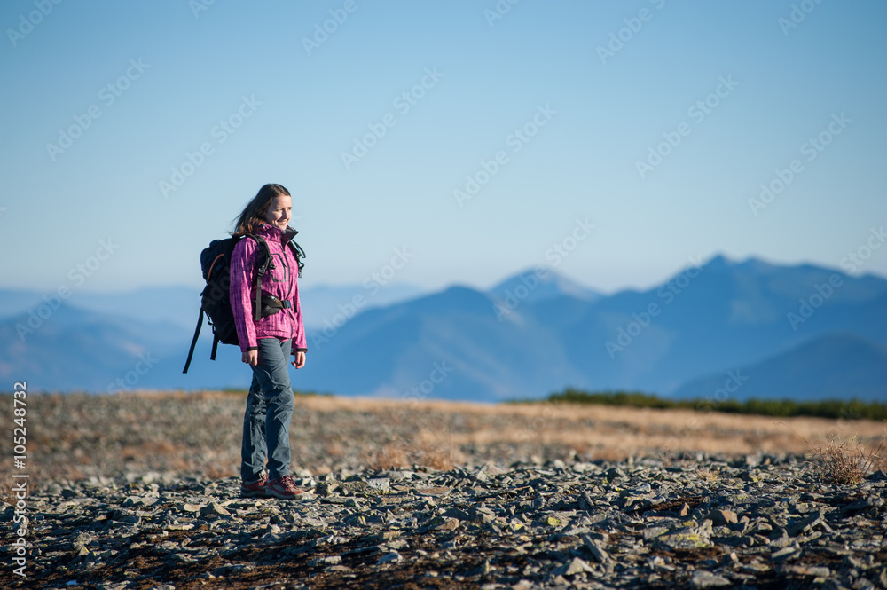 Young beautiful female hiker standing on the rocky mountain plato and enjoying beautiful nature landscape. Blue mountains and sky on background. Woman wearing pink jacket with backpack on.
