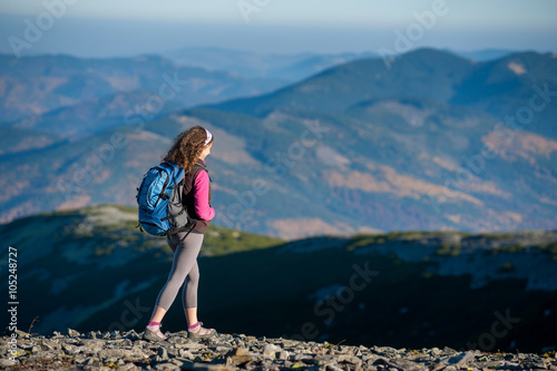 Young woman tourist is climbing down on mountain plato with big backpack on. Beautiful mountains on background. Sunny autumn day.