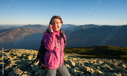 Portrait of young happy woman backpacker standing on rocky top of the mountain, smiling, looking to the distance and talking cell phone. Side view. Healthy lifestyle concept. Sunny autumn evening.
