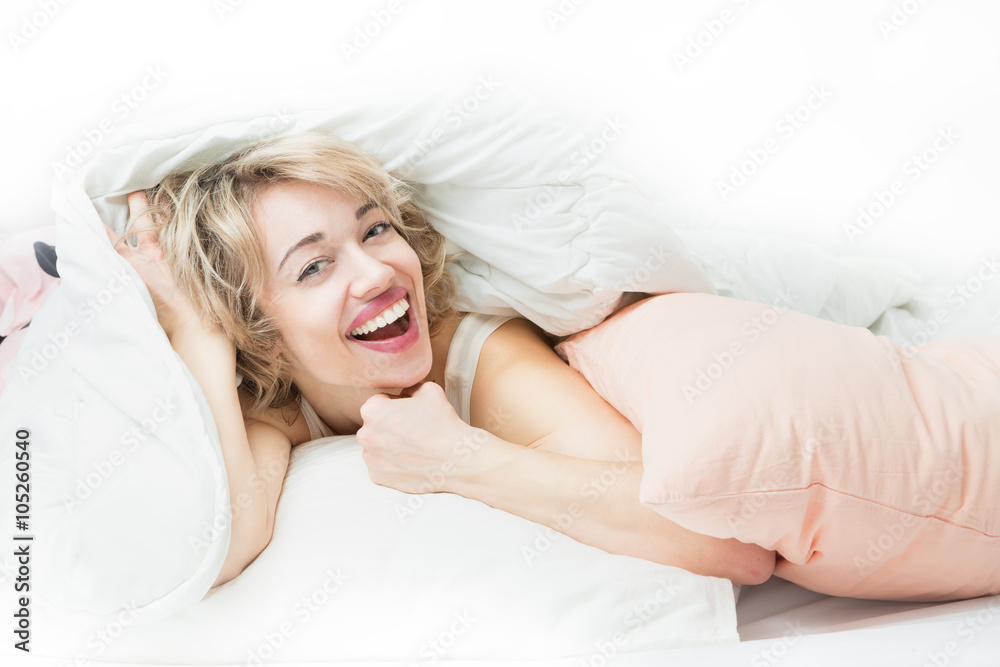 Laughing happy woman peeking out from under the blanket. Wide sincere beautiful smile. Linen pillows and duvet Laughing happy woman peeking out from under the blanket. Wide sincere beautiful smile. Linen pillows and duvet