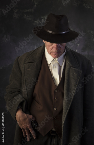 Sinister/Low key image of man in overcoat & hat in sinister pose with hand on gun against portrait background