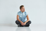 Brutal man in a shirt with short sleeves sitting in lotus posture, white background and floor, looking away