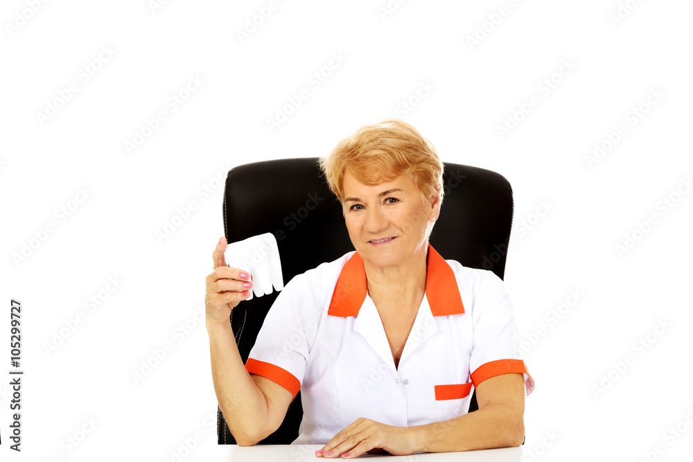 Smile elderly female dentist sitting behind the desk and holds big tooth model