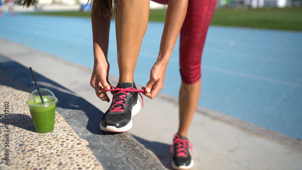 Athlete runner getting ready for run with green smoothie tying running shoes on outdoor athletic track and field with runners in the background. Legs and feet closeup. Athlete runner getting ready for run with green smoothie tying running shoes on outdoor athletic track and field with runners in the background. Legs and feet closeup.