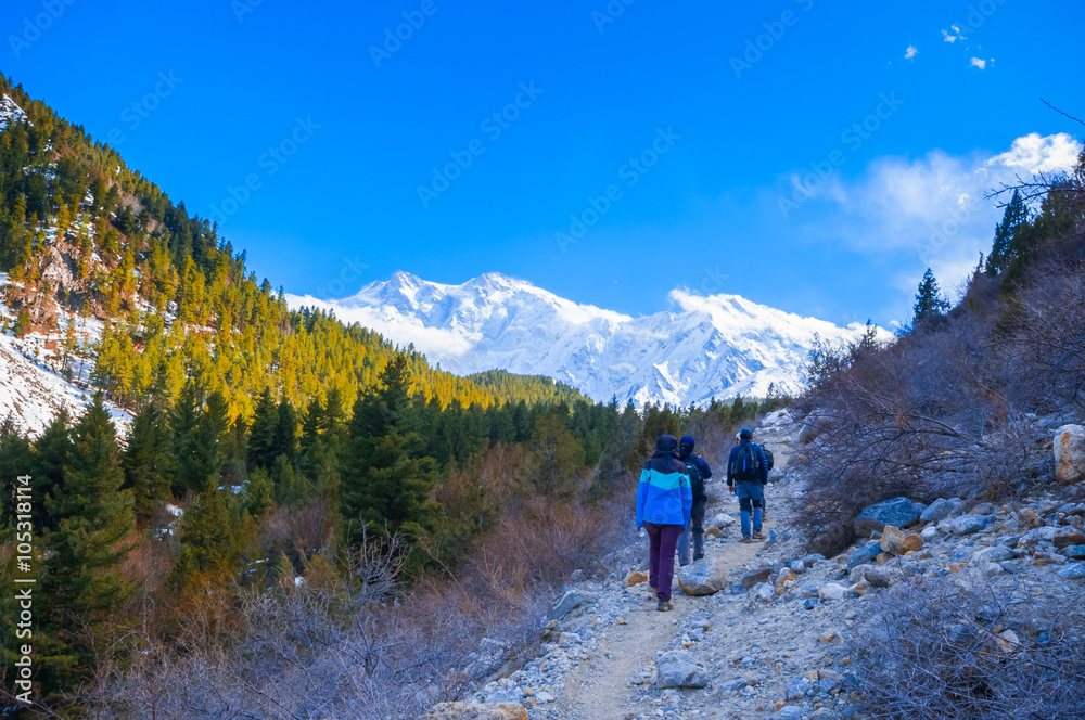 people with backpacks walking along the road. There are mountains on the horizon. The sky is blue and cloudy.