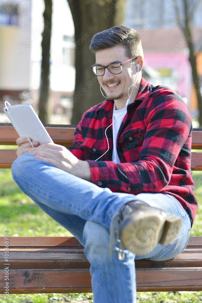 Obraz premium Portrait of young man looking at the camera and holding tablet PC. Happy man in casual working in the park while sitting on the bench.