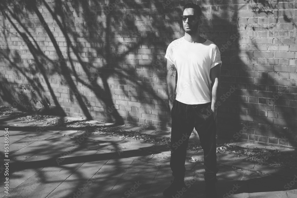Fototapeta premium Photo bearded man with tattoo wearing blank white tshirt, sunglasses. Stands in front of a brick wall. City street background. Horizontal mockup. Black, White