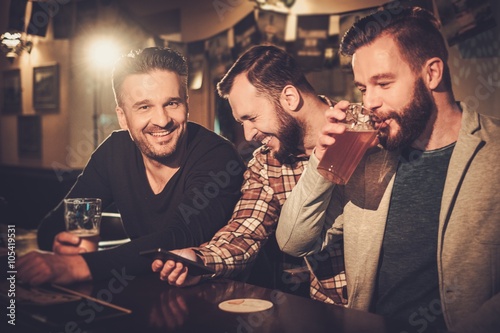 Wall Mural Cheerful old friends having fun with smartphone and drinking draft beer at bar counter in pub