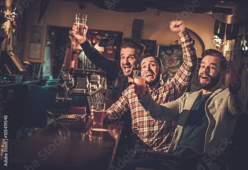 Photography Cheerful old friends having fun watching a football game on TV and drinking draft beer at bar counter in pub